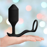 An open palm showcases the B-Vibe Vibrating Cock Ring - Snug & Tug (XL), a sleek black silicone design with a practical looped handle. Set against a backdrop of pastel bokeh lights that create a gentle ambiance, this weighted accessory enhances sensations and provides an elegant yet comfortable experience.